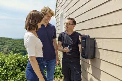 Uśmiechnięte młode małżeństwo i ekspert Hager stojący przy zamontowanej na ścianie domu stacji witty one w trakcie prezentacji produktu