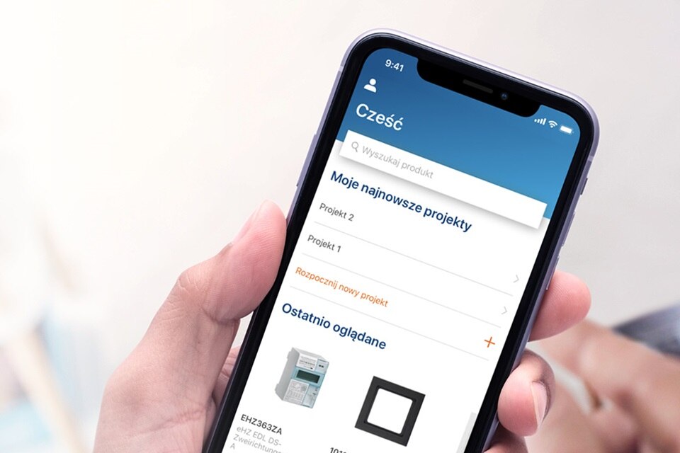 Ręka trzymająca smartfon z interfejsem mobilnej aplikacji Hager w języku polskim, wyświetlająca listę projektów i produkty do rozdziału energii elektrycznej.