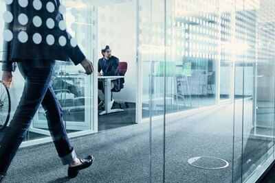Osoba przechodząca przez nowoczesne biuro z przeszklonymi ścianami i druga osoba pracująca w środku sali konferencyjnej