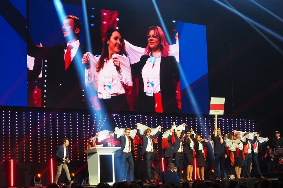 Grupa osób na scenie podczas wydarzenia z dużymi ekranami wyświetlającymi ich wizerunki oraz tabliczką z napisem 'Polska'