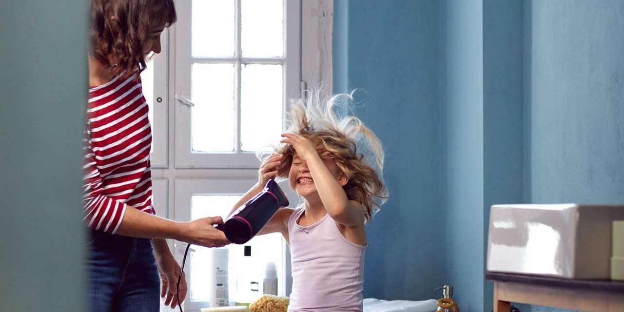 Skrattande flicka på badkarskanten får sitt hår torkat med en hårfön av sin mamma