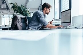 En man med glasögon och blå skjorta sitter vid ett skrivbord och planerar sitt arbete vid sin laptop