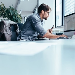 En man med glasögon och blå skjorta sitter vid ett skrivbord och planerar sitt arbete vid sin laptop