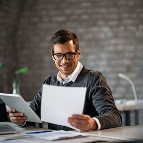 Man sitter vid sitt skrivbord framför sin laptop och läser från ett papper