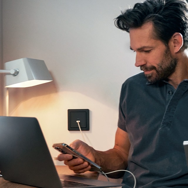 En person använder en smartphone som är ansluten till ett Hager vägguttag med usb bredvid en skrivbordslampa och en bärbar dator.